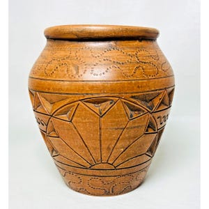 May include: A brown, handcrafted pottery vase with intricate carved geometric and floral patterns. The vase has a wide, rounded body and a slightly flared rim. The design includes sunburst and dotted line details.
