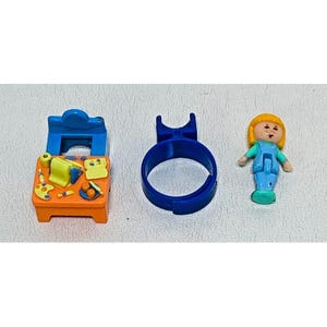 May include: A collection of miniature toys, including a blue desk with art supplies, a blue ring, and a small doll with yellow hair and blue clothing. The desk is orange and has a blue chair attached.