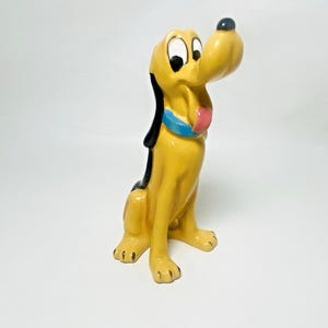 May include: A ceramic figurine of Pluto, a yellow dog with black ears and a blue collar. The dog has a pink tongue sticking out and is sitting upright. The figurine is a collectible item.