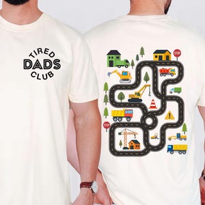 May include: Ivory-colored t-shirt with the text "TIRED DADS CLUB" on the front. The back features a colorful road map design with construction vehicles, houses, and trees. The shirt is made of Comfort Colors 1717 material.