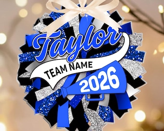 May include: A round cheerleading pom-pom ornament featuring blue, black, and silver glitter strips. It includes a white banner with the text Taylor, Team Name, and 2026, along with a blue megaphone graphic and a cream-colored ribbon for hanging.
