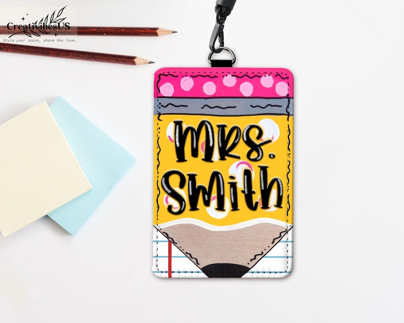 K&ouml;nnte beinhalten: Ein gelber, stiftf&ouml;rmiger Ausweishalter mit dem Text "MRS. SMITH" in Schwarz. Die Oberseite hat ein rosa Tupfenmuster und die Unterseite ein Bleistiftspitzen-Design. Enth&auml;lt ein schwarzes Schl&uuml;sselband und befindet sich neben Bleistiften und Haftnotizen.
