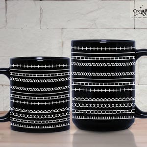 May include: Two black ceramic mugs with white stitched-style patterns. The mugs have a glossy finish and large handles. The patterns include horizontal lines of dashes, X's, and other decorative stitches. The mugs are on a wooden surface.