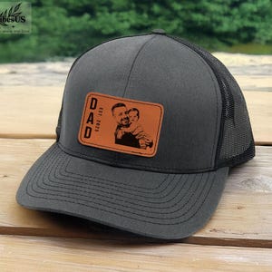 May include: A black and grey trucker hat with a brown leather patch that says "DAD EST 2025" and has a photo of a man and a child on it.