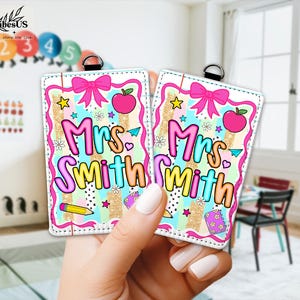 May include: Two rectangular teacher name tags with a colorful design. Each tag features the name "Mrs. Smith" in a rainbow font, a pink bow, an apple, and school-themed graphics. The tags are held by a hand.