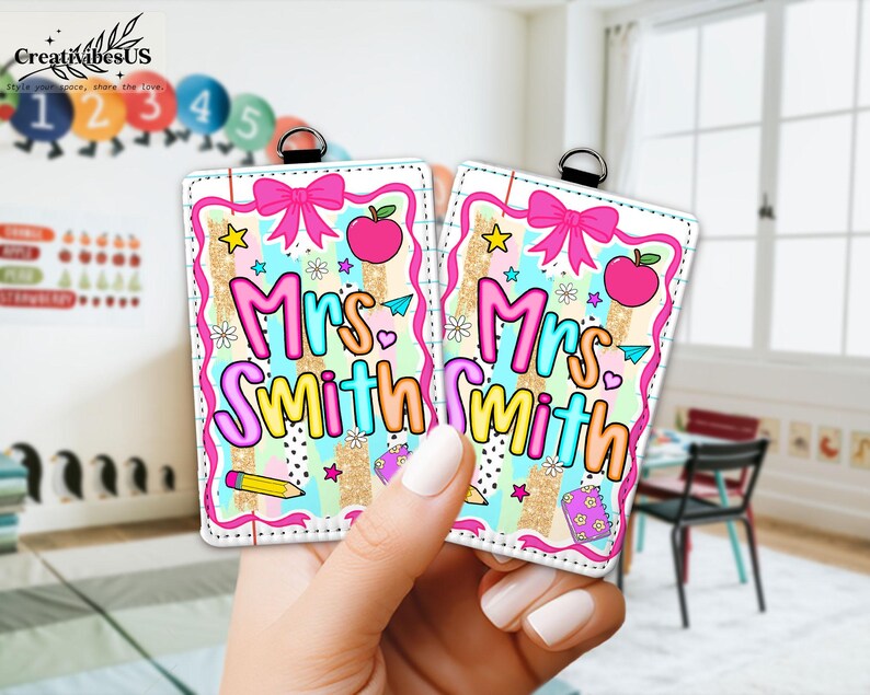 May include: Two rectangular teacher name tags with a colorful design. Each tag features the name "Mrs. Smith" in a rainbow font, a pink bow, an apple, and school-themed graphics. The tags are held by a hand.