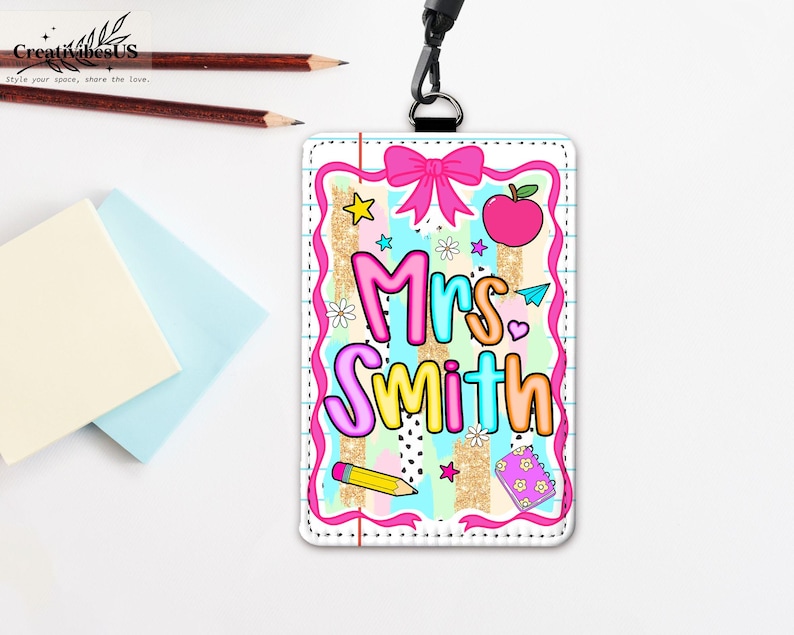May include: A colorful teacher ID badge with the name "Mrs. Smith" in bubble letters. The badge features a pink bow, apple, stars, pencil, and notebook designs. It has a black lanyard and is on a white surface.