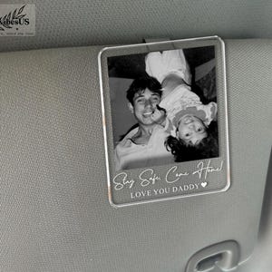K&ouml;nnte beinhalten: Ein rechteckiger, durchsichtiger Acryl-Clip f&uuml;r die Sonnenblende im Auto mit einem Schwarz-Wei&szlig;-Foto eines Erwachsenen und eines Kindes. Der Text "Stay Safe, Come Home!" und "LOVE YOU DADDY" mit einem kleinen Herzsymbol ist unten wei&szlig; aufgedruckt.