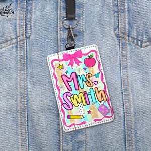 May include: A colorful teacher ID badge with the name "Mrs. Smith" in bubble letters. The badge features a pink bow, an apple, stars, and a pencil. It hangs from a black lanyard attached to a silver clip.