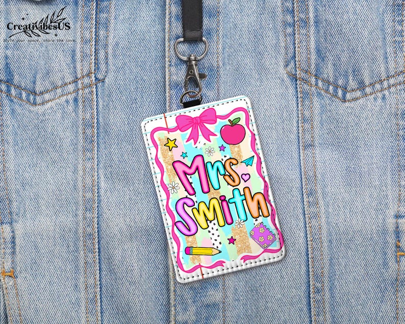 May include: A colorful teacher ID badge with the name "Mrs. Smith" in bubble letters. The badge features a pink bow, an apple, stars, and a pencil. It hangs from a black lanyard attached to a silver clip.