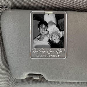 K&ouml;nnte beinhalten: Ein rechteckiger, transparenter Acryl-Clip f&uuml;r die Sonnenblende mit einem schwarz-wei&szlig;en Foto einer Person, die ein Kind h&auml;lt. Der Text "Stay Safe, Come Home!" und "LOVE YOU DADDY" mit einem Herzsymbol sind unten in Wei&szlig; aufgedruckt.