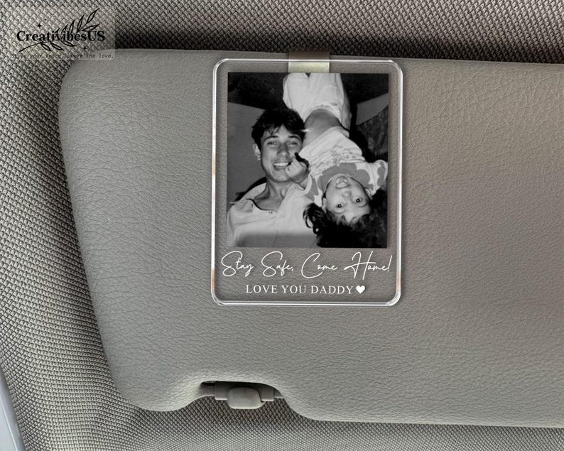K&ouml;nnte beinhalten: Ein rechteckiger, transparenter Acryl-Clip f&uuml;r die Sonnenblende mit einem schwarz-wei&szlig;en Foto einer Person, die ein Kind h&auml;lt. Der Text "Stay Safe, Come Home!" und "LOVE YOU DADDY" mit einem Herzsymbol sind unten in Wei&szlig; aufgedruckt.