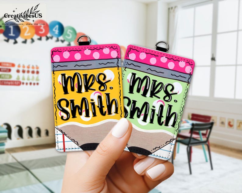 K&ouml;nnte beinhalten: Zwei bleistiftf&ouml;rmige Schl&uuml;sselanh&auml;nger, einer gelb und einer gr&uuml;n, werden in einer Hand gehalten. Jeder Schl&uuml;sselanh&auml;nger tr&auml;gt den Namen "Mrs. Smith" in schwarzen Buchstaben. Die Oberseite jedes Schl&uuml;sselanh&auml;ngers ist rosa mit wei&szlig;en Punkten.