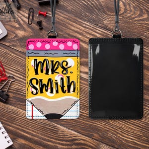 K&ouml;nnte beinhalten: Ein personalisierter Lehrer-Ausweishalter mit Bleistift-Design. Der Ausweishalter tr&auml;gt den Namen "MRS. SMITH" in fetten, schwarzen Buchstaben auf gelbem Hintergrund. Das Design umfasst rosa Punkte, Grau und eine Bleistiftspitze. Ein schwarzes Schl&uuml;sselband ist befestigt.