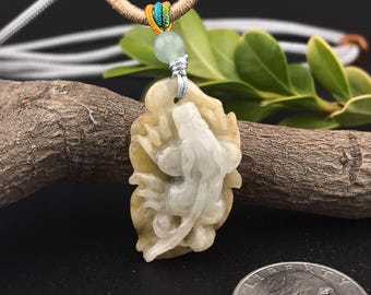 May include: A carved jade pendant with a detailed floral and figure design. The pendant is a mix of light green and tan colors, suspended on a beige and gray cord. The background includes a wooden branch and green leaves.