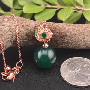 May include: A rose gold necklace with a pendant featuring a green gemstone sphere. The pendant is adorned with a gold-colored lotus flower design, with a small green gem at its center. The necklace is displayed on a wooden branch.