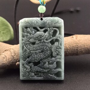 May include: A jade pendant necklace featuring a detailed carving of a dragon. The pendant is a dark green color and has a rectangular shape. The necklace is made of black cord and has a green bead at the top.