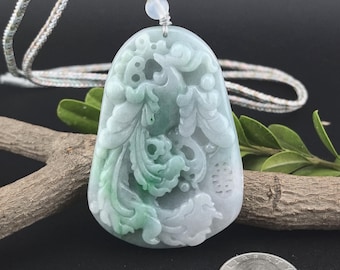 May include: A carved jade pendant with a detailed floral and bird design in shades of green and white. The pendant is suspended from a woven cord. The jade is approximately 2 inches in length.