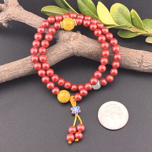 May include: Red beaded bracelet with two yellow decorative beads and a small blue and white flower charm. The bracelet is draped over a wooden branch with green leaves. A silver coin is next to the bracelet.