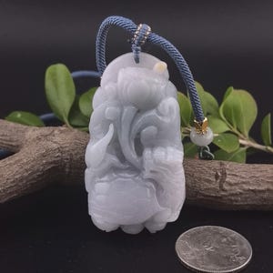 May include: A carved jade pendant in shades of pale green and white, featuring a floral design. The pendant is suspended from a blue cord with gold accents. A coin rests below the pendant.