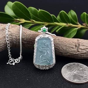 May include: A silver necklace with a rectangular, light blue jade pendant. The pendant features a carved design and is surrounded by a border of small, clear gemstones. A small green gemstone is at the top of the pendant. A US quarter is in the image.