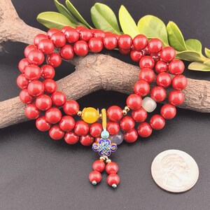 May include: A red beaded bracelet with a decorative charm. The bracelet is made of round, glossy red beads. A yellow bead and a decorative charm with blue and gold accents are also present. A US quarter is next to the bracelet.