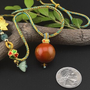 May include: A necklace with a large, round, reddish-brown pendant. The necklace cord is gold and teal. A quarter coin is next to the pendant for size comparison. The necklace also has a gold charm with a green and red clover.