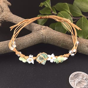 May include: A green fluorite crystal charm bracelet with white flower beads and brown cord. The bracelet is tied with a knot and is about the size of a US quarter.