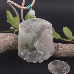 May include: A jade pendant with a carved design of a mythical creature. The pendant is a light green color with hints of purple and is suspended from a beige cord. A small, round, light green bead accents the cord. A coin is in the foreground.