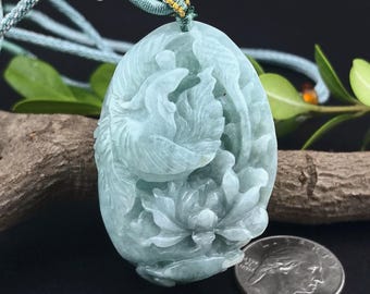 May include: A carved jade pendant in shades of pale green, featuring a bird and floral design. The pendant is suspended from a light blue cord with gold accents. A silver coin is next to the pendant.