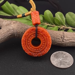 May include: A reddish-orange pendant necklace with intricate carved patterns. The pendant is round with a central hole and is suspended from a black cord with a decorative gold and black woven section. A U.S. quarter is next to the pendant.