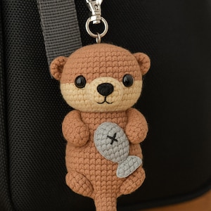 Op de afbeelding: Een handgemaakte gehaakte otter sleutelhanger in bruin- en beigetinten, met een grijze vis. De otter heeft zwarte ogen en een zwarte neus. De sleutelhanger is bevestigd aan een zilverkleurige sluiting.