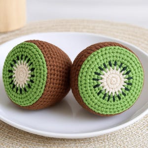 May include: Two crocheted kiwi fruit halves. Each has a brown exterior, a green rind, and a cream-coloured centre with black seed details. The kiwis are presented on a white plate.