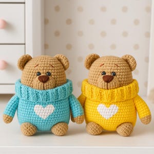 May include: Two handmade crochet teddy bears. One bear wears a blue sweater, the other a yellow sweater. Each bear has a white heart on the front. The bears have brown noses and eyes, and are sitting on a white surface.