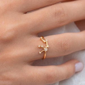 May include: A gold ring with a constellation design featuring small clear stones. The ring is worn on a finger.