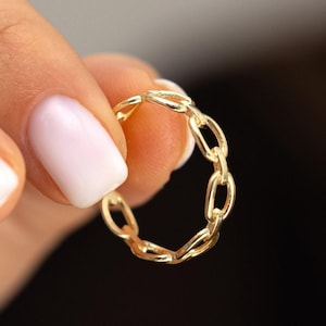 May include: A gold chain ring with a delicate, linked design. The ring is held in a hand with white nail polish.