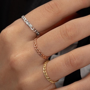 May include: Three gold chain rings stacked on a finger. The rings are different sizes and are made of a thick, chunky chain.