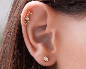 May include: A close-up of a person's ear with two gold earrings. The earring on the upper ear is a curved bar with a constellation of small stars. The earring on the lower ear is a single star.