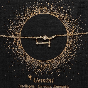 Puede incluir: Collar de oro con un dise&ntilde;o de constelaci&oacute;n que representa el signo zodiacal G&eacute;minis. El collar est&aacute; sobre un fondo negro con purpurina dorada. El texto "G&eacute;minis" e "Inteligente, Curioso, En&eacute;rgico" est&aacute; impreso debajo del collar.
