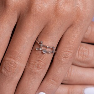 May include: A silver ring with a constellation design featuring small clear stones. The ring is worn on a person's finger.