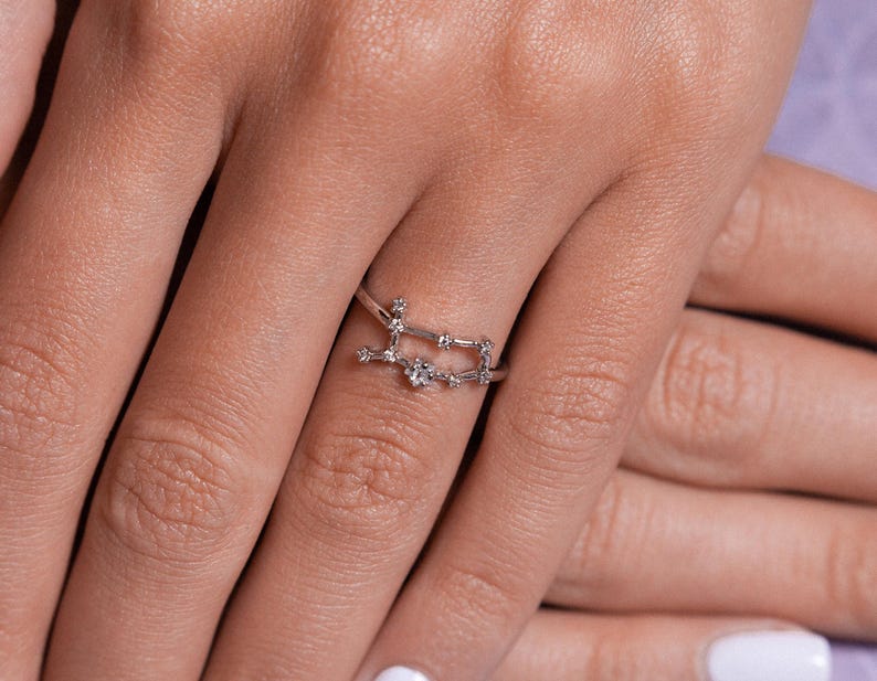 May include: A silver ring with a constellation design featuring small clear stones. The ring is worn on a person's finger.