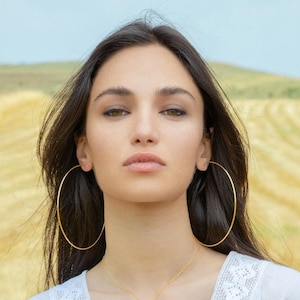 Peut inclure: Une femme aux longs cheveux noirs portant de grandes boucles d'oreilles en or. Elle porte un haut blanc et un collier en or.