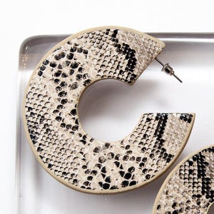 Puede incluir: Un par de pendientes grandes de aro con estampado de piel de serpiente. Los pendientes son de color beige con detalles negros.