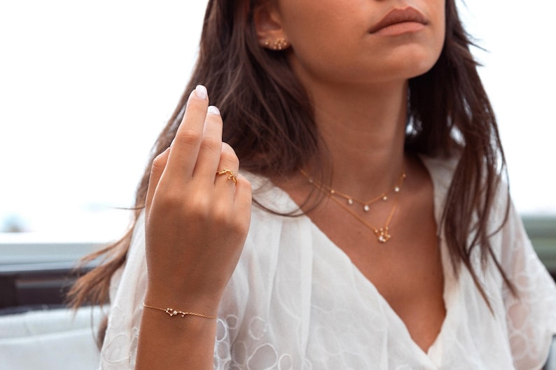 May include: A woman wearing a white top with a gold necklace and bracelet. She is also wearing a gold ring on her finger.