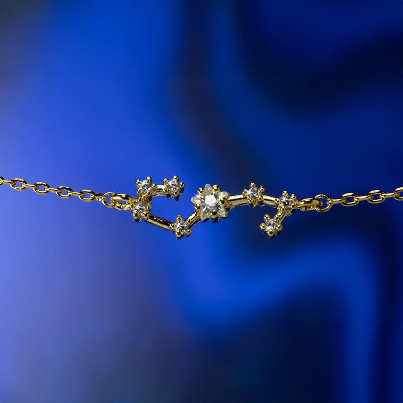 Puede incluir: Un collar de cadena dorada con un colgante de constelaci&oacute;n con peque&ntilde;as piedras transparentes. El colgante tiene la forma de la constelaci&oacute;n de Acuario.