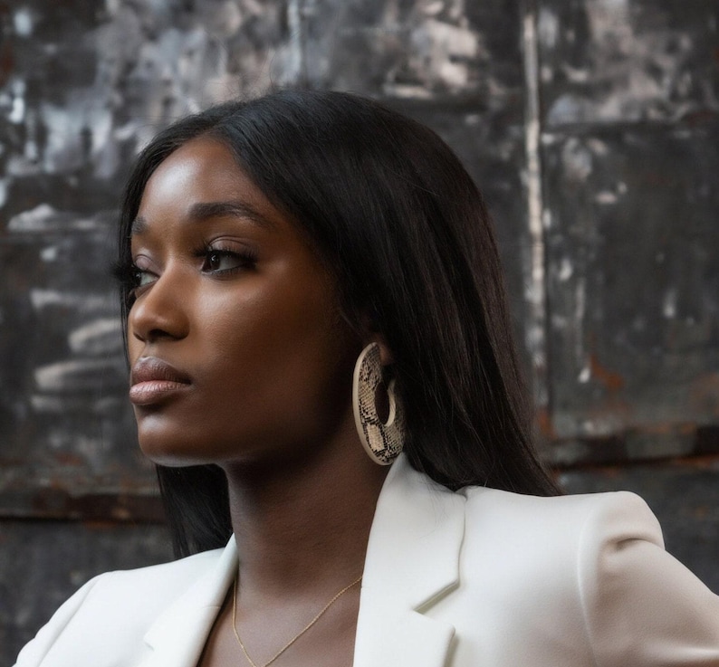 Puede incluir: Una mujer con cabello casta&ntilde;o oscuro que lleva una chaqueta blanca y un par de pendientes grandes, redondos, con estampado de piel de serpiente.