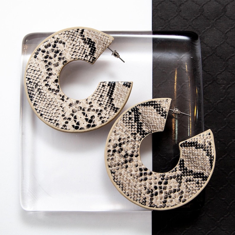 Puede incluir: Un par de pendientes grandes de aro con estampado de piel de serpiente. Los pendientes son beige con marcas negras.