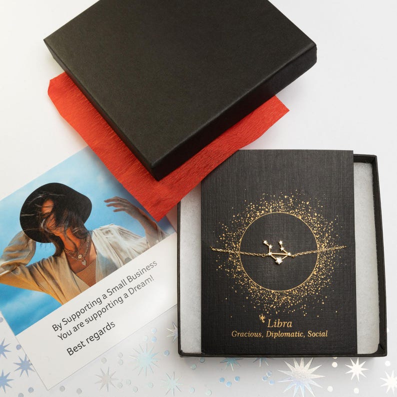 Puede incluir: Una caja negra con un forro de papel de seda rojo, que contiene una pulsera de oro con un dije de constelaci&oacute;n de Libra. La caja tiene una tarjeta blanca con el texto "Libra, Gracioso, Diplom&aacute;tico, Social" y un mensaje de agradecimiento.
