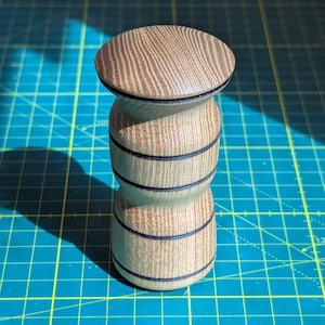 Peut inclure: Un bouchon de bouteille en bois avec un grain de bois brun clair et des accents noirs. Le bouchon a un dessus arrondi et un corps cylindrique avec trois bandes noires.