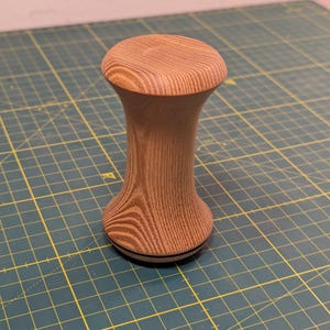 Könnte beinhalten: Ein Holztamper mit braunem und weißem Holzmaserungsmuster. Der Tamper hat eine abgerundete Oberseite und eine leicht ausgestellte Basis.